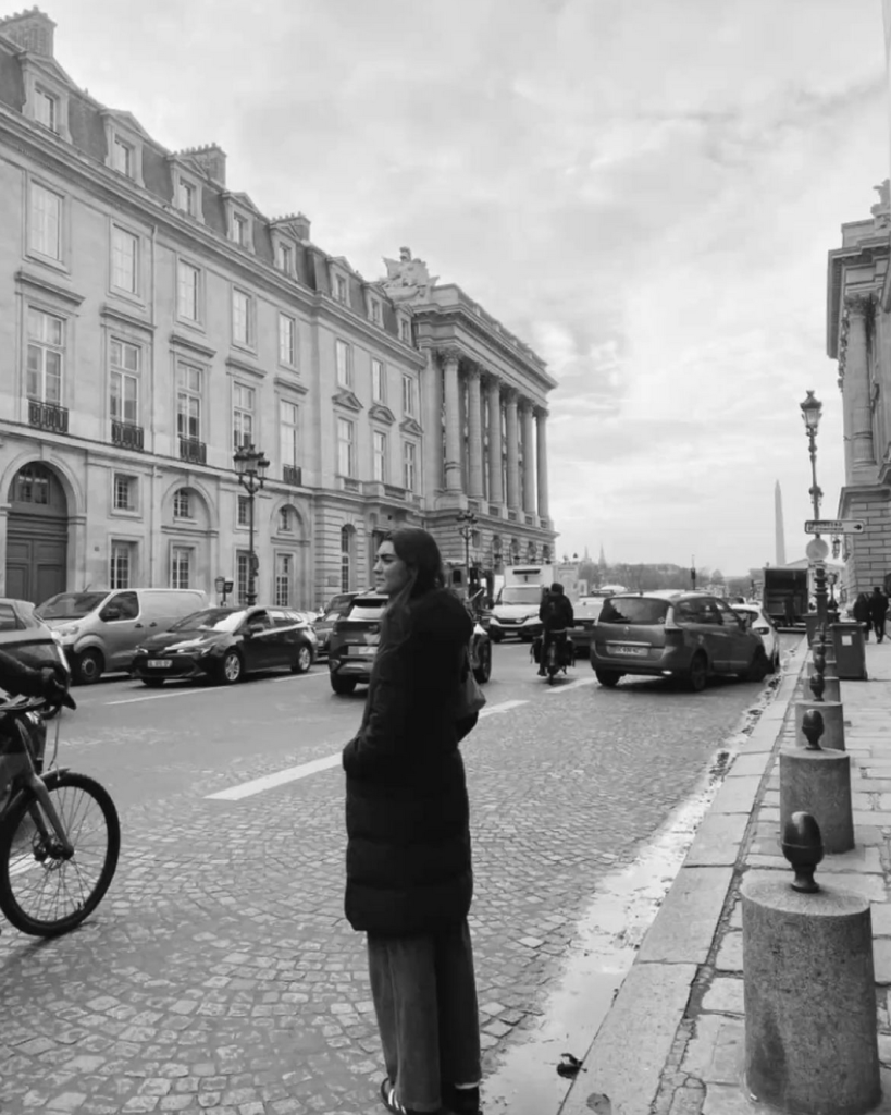 A girl walking around a roman city about to cross the street wearing a long trench coat.